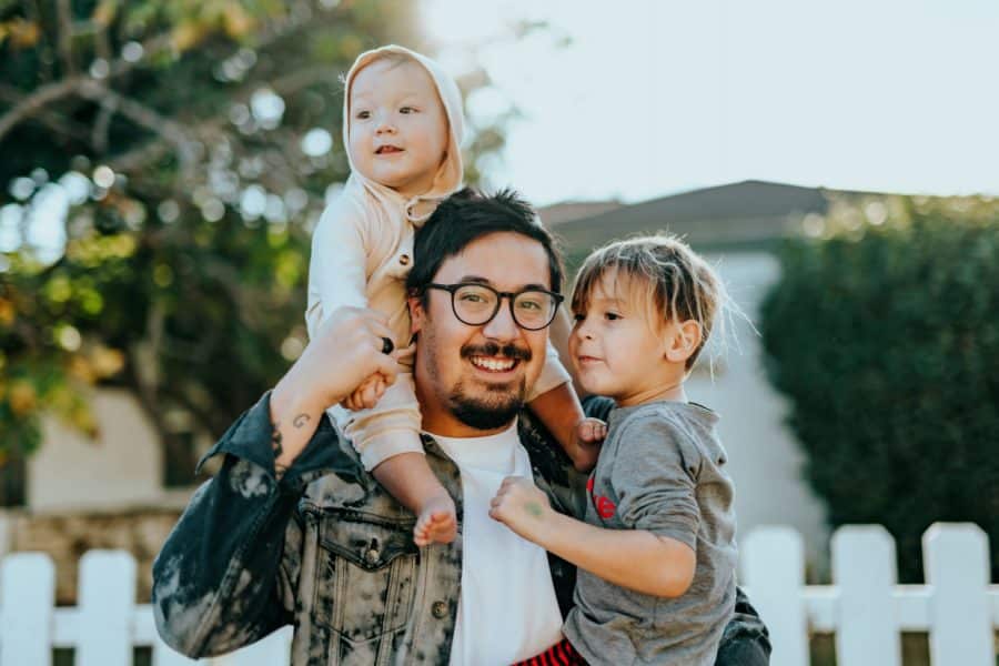 Father smiling while holding a toddler on his shoulders and a young child beside him, outdoors with greenery and a white fence in the background, symbolizing family and home.