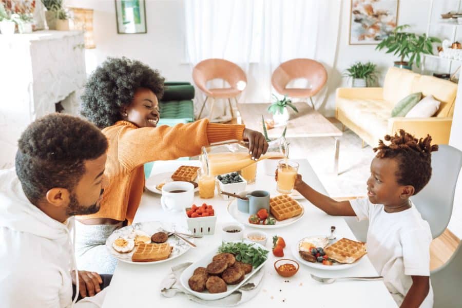 Family enjoying breakfast at a dining table with waffles, fruit, and drinks in a bright, cozy kitchen setting.