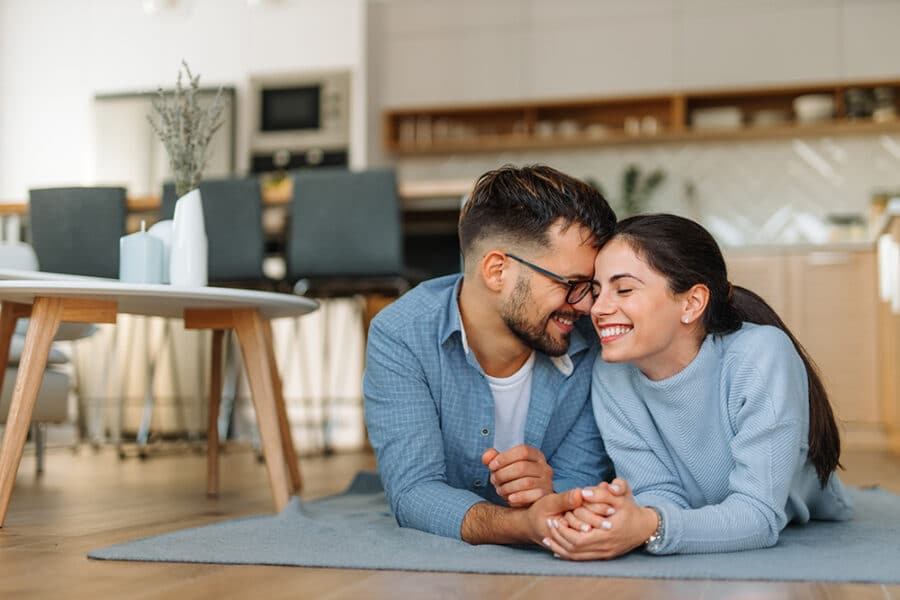 Couple smiling and holding hands on a rug in a modern kitchen setting, reflecting home buying and relationship themes.